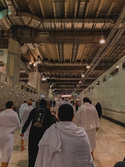 A group of people wearing white garments walk through a large, spacious corridor under a high industrial-style ceiling with exposed ducts and lights. The floor is tiled, and the walls are lined with glass panels. The environment suggests a peaceful and solemn atmosphere as the individuals proceed forward.