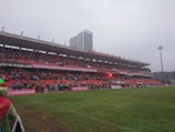 A sports stadium with a large crowd of spectators wearing red seated in the stands. The field is green and wet, suggesting a recent or ongoing rain. Banners with text are displayed above the audience. In the background, a tall building is visible, and the sky is overcast.