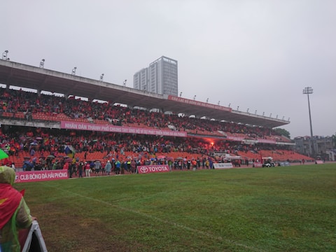 A sports stadium with a large crowd of spectators wearing red seated in the stands. The field is green and wet, suggesting a recent or ongoing rain. Banners with text are displayed above the audience. In the background, a tall building is visible, and the sky is overcast.