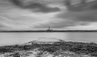 A black and white seascape with a calm body of water extending into the distance. An oil rig is visible on the horizon, under a sky with thick, dramatic clouds. The foreground consists of a rocky shore with seaweed scattered along the ground.