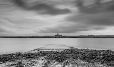 A black and white seascape with a calm body of water extending into the distance. An oil rig is visible on the horizon, under a sky with thick, dramatic clouds. The foreground consists of a rocky shore with seaweed scattered along the ground.