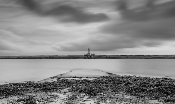 A black and white seascape with a calm body of water extending into the distance. An oil rig is visible on the horizon, under a sky with thick, dramatic clouds. The foreground consists of a rocky shore with seaweed scattered along the ground.