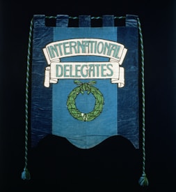 A blue fabric banner featuring a design with the words 'International Delegates' in white lettering. Below the text, there is a green laurel wreath. Decorative twisted ropes with tassels hang from both sides.