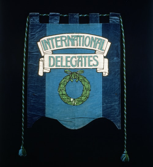 A blue fabric banner featuring a design with the words 'International Delegates' in white lettering. Below the text, there is a green laurel wreath. Decorative twisted ropes with tassels hang from both sides.