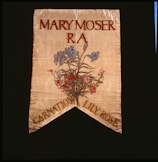 A decorative banner featuring the name Mary Moser with R A beneath it. Below the text, there is an artistic depiction of a floral arrangement featuring blue, pink, and red flowers. The banner has a pointed end with the words 'Carnation Lily Rose' at the bottom.