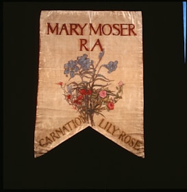 A decorative banner featuring the name Mary Moser with R A beneath it. Below the text, there is an artistic depiction of a floral arrangement featuring blue, pink, and red flowers. The banner has a pointed end with the words 'Carnation Lily Rose' at the bottom.