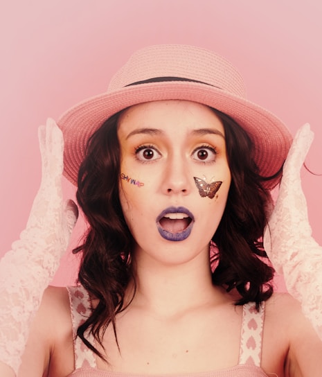 A person with an expression of surprise or wonder wears a pink hat and lace gloves. The face has colorful makeup featuring a butterfly and playful text. The background is a soft pink, complementing the overall color scheme.