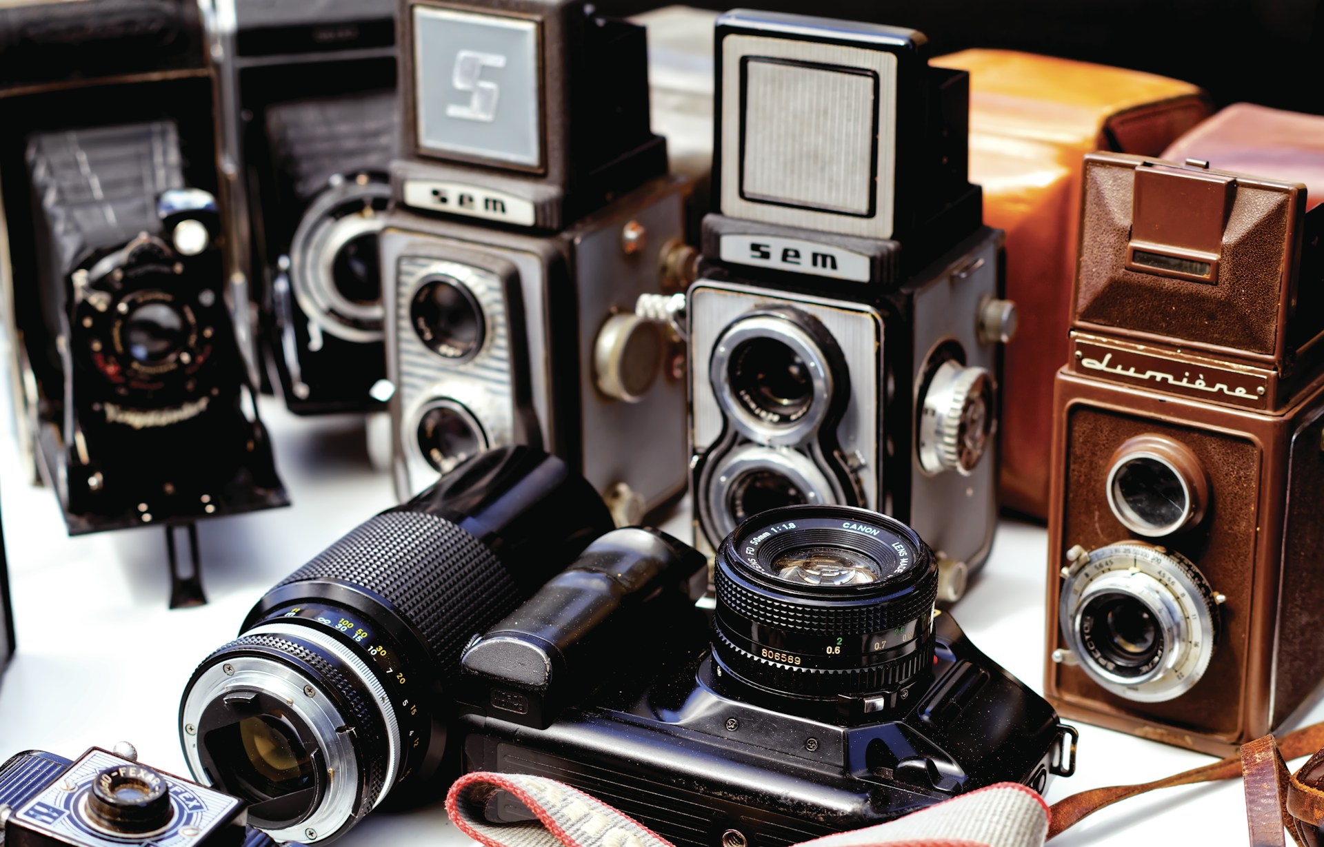 A collection of vintage cameras is arranged, showcasing a variety of styles and designs. The cameras feature different lens sizes and types, reflecting an era of classic photography. Some cameras are box-shaped, while others have a more rectangular build. The materials appear to range from metal to leather finishes, adding to the nostalgic feel.