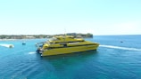 A large, yellow ferry named 'Bounty Cruises' sails on a clear blue sea, with a shoreline visible in the background. The water is calm and other small boats are seen nearby. The horizon shows a mix of vibrant greenery and buildings along the coastline.