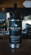 A large black and silver coffee dispenser with the branding 'True Stone Coffee Roasters' displayed prominently. Below the logo, the phrases 'Fresh. Local. Independent.' and 'Fresh Coffee. Cool Beans.' are printed. In the background, a blurred coffee shop setting with dark cabinets and various equipment is visible.