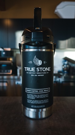 A large black and silver coffee dispenser with the branding 'True Stone Coffee Roasters' displayed prominently. Below the logo, the phrases 'Fresh. Local. Independent.' and 'Fresh Coffee. Cool Beans.' are printed. In the background, a blurred coffee shop setting with dark cabinets and various equipment is visible.