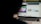A partially obscured view of a laptop screen displaying two separate browser tabs. The left tab shows a children's cartoon video, while the right tab appears to be a webpage with photos and metadata, possibly related to stock photography. The foreground is dark, highlighting the screen's content.