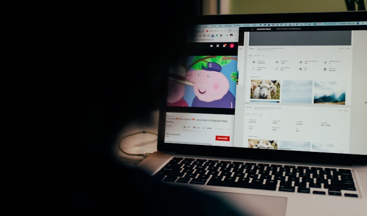 A partially obscured view of a laptop screen displaying two separate browser tabs. The left tab shows a children's cartoon video, while the right tab appears to be a webpage with photos and metadata, possibly related to stock photography. The foreground is dark, highlighting the screen's content.