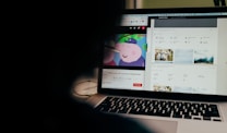 A partially obscured view of a laptop screen displaying two separate browser tabs. The left tab shows a children's cartoon video, while the right tab appears to be a webpage with photos and metadata, possibly related to stock photography. The foreground is dark, highlighting the screen's content.