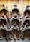 A well-organized display of horse saddles mounted on a wooden wall, neatly arranged in rows. Each saddle is made of brown leather with distinct textures and includes straps and other accessories. The setup suggests a focus on equestrian themes or a stable environment.