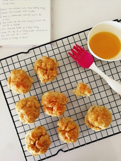 Several freshly baked biscuits are cooling on a black wire rack. A pink pastry brush is situated next to them, along with a bowl containing a golden liquid, likely honey or butter. In the background is an open notebook with a recipe written on it.