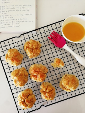 Several freshly baked biscuits are cooling on a black wire rack. A pink pastry brush is situated next to them, along with a bowl containing a golden liquid, likely honey or butter. In the background is an open notebook with a recipe written on it.