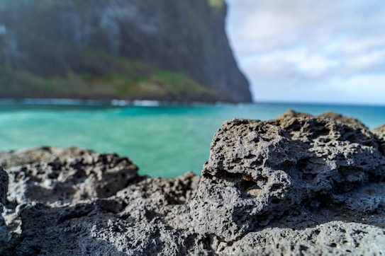 A close-up of rugged, dark volcanic rock with a blurred background showcasing a turquoise ocean and a large cliff under a partly cloudy sky.