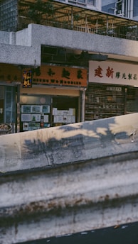 A city street view featuring storefronts with signage in a mix of English and Chinese characters. A large, weathered concrete barrier occupies the foreground, casting shadows from trees. Above the shops, greenery is visible on a balcony area with a lattice framework.
