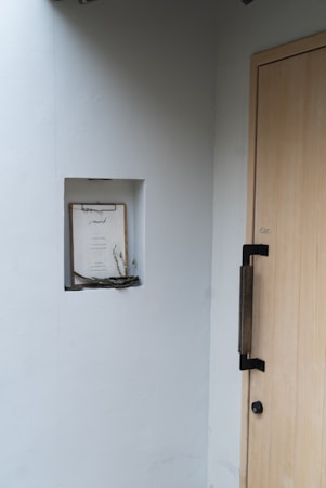 A minimalistic entryway featuring a light wooden door with a black handle. On the adjacent wall, a small recessed shelf holds a clipboard with a paper and some dried twigs. The wall is painted in a soft white color, providing a clean and simple aesthetic.