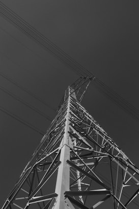 A tall electricity transmission tower is captured from a low angle, displaying its intricate metal framework against a dark sky. Several power lines extend outward from the tower's peak, emphasizing its height and structure.