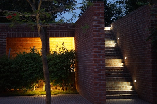A brick structure features a small outdoor stairway with integrated lighting. Soft yellow illumination lights up the foliage below, casting shadows against the brick walls. The ambiance suggests a quiet evening atmosphere with natural elements like trees blending with the architectural design.