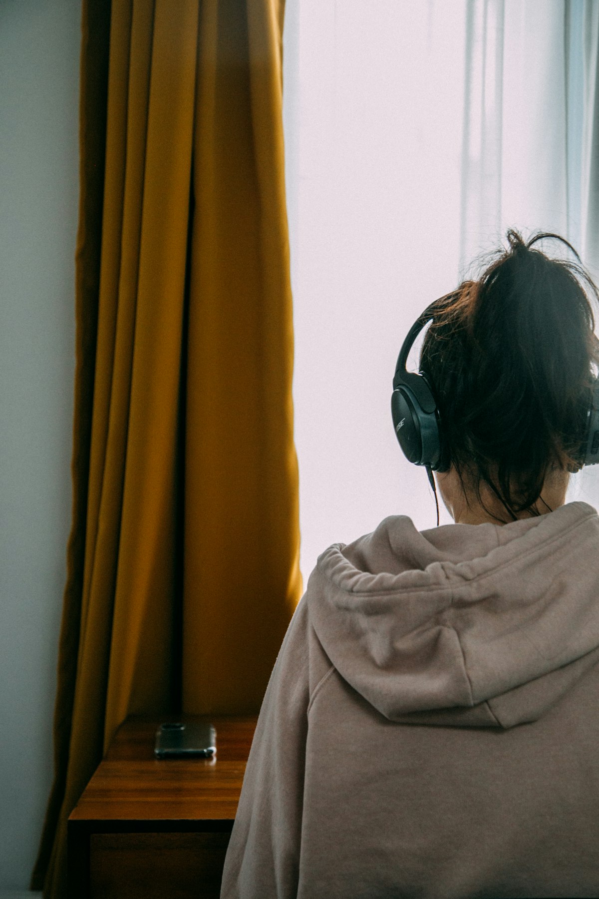A person seen from behind in a hoodie and black over-ear headphones, facing a window in warm light, deep-work register
