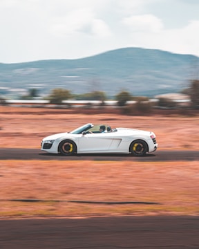 A sleek white sports car speeds along an open road with blurred motion, suggesting high speed. The background features a scenic view with hills and partially cloudy skies. The driver is wearing a helmet, indicating a focus on safety and possibly a race setting.