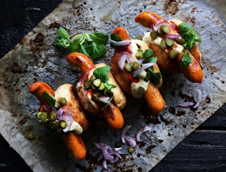 Three grilled sausages are placed in buns on a baking sheet. They are garnished with green leafy herbs, slices of red onion, and small pickles. A creamy sauce is drizzled on top, with a sprinkle of black pepper for seasoning.