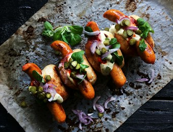 Three grilled sausages are placed in buns on a baking sheet. They are garnished with green leafy herbs, slices of red onion, and small pickles. A creamy sauce is drizzled on top, with a sprinkle of black pepper for seasoning.