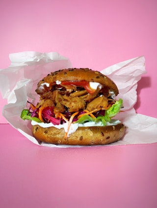 A deliciously presented burger with a seeded bun, layered with fresh lettuce, vibrant coleslaw, mayonnaise, and savory fillings like sliced chicken or tofu. The background is a solid pink, creating a striking contrast with the burger's colorful ingredients. The burger is partially wrapped in white parchment paper.