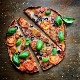 A delicious pizza with a thin crust, topped with tomato slices, pepperoni, black olives, fresh basil leaves, and melted cheese. The pizza is cut into large slices and sprinkled with herbs, set on a rustic wooden surface.