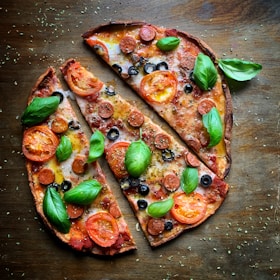 A delicious pizza with a thin crust, topped with tomato slices, pepperoni, black olives, fresh basil leaves, and melted cheese. The pizza is cut into large slices and sprinkled with herbs, set on a rustic wooden surface.