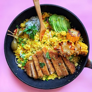 A vibrant dish is presented in a black pan, featuring golden couscous mixed with fresh herbs and vegetables. The top is adorned with sliced avocado, charred corn on the cob, caperberries, and crispy breaded cutlets. A wooden spoon is placed in the middle, enhancing the composition with a touch of rustic charm.