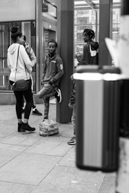 A group of young people are gathered in what appears to be an urban setting, possibly a bus or train station. One person leans against a post with a relaxed posture, while others are standing nearby engaged in conversation. There is a backpack placed on the ground, and the scene is captured in black and white.