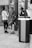 A group of young people are gathered in what appears to be an urban setting, possibly a bus or train station. One person leans against a post with a relaxed posture, while others are standing nearby engaged in conversation. There is a backpack placed on the ground, and the scene is captured in black and white.