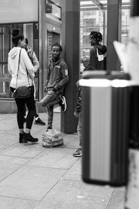 A group of young people are gathered in what appears to be an urban setting, possibly a bus or train station. One person leans against a post with a relaxed posture, while others are standing nearby engaged in conversation. There is a backpack placed on the ground, and the scene is captured in black and white.