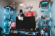 A collection of action figures and collectibles is displayed on glass shelves, with a central large flat-screen TV. Star Wars characters, including Darth Vader and Stormtroopers, are prominent, while various other figures and memorabilia fill the rest of the space. The setup includes bright blue LED lighting.