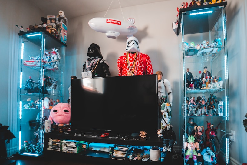 A collection of action figures and collectibles is displayed on glass shelves, with a central large flat-screen TV. Star Wars characters, including Darth Vader and Stormtroopers, are prominent, while various other figures and memorabilia fill the rest of the space. The setup includes bright blue LED lighting.