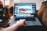 A person's hand rests on a laptop keyboard while the screen displays an image editing software with a photo of parked cars. The setting appears casual, possibly in a living room with soft focus furniture in the background.