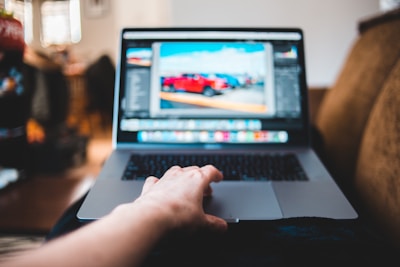 A person's hand rests on a laptop keyboard while the screen displays an image editing software with a photo of parked cars. The setting appears casual, possibly in a living room with soft focus furniture in the background.