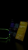 The image features a dimly lit room with two electronic devices visible. One device displays a screen with a music app playing, showing lyrics in yellow against a black backdrop. Another device, possibly a speaker, has a green light outline. The overall lighting is low, emphasizing the glow from the screen and lights.