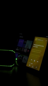 The image features a dimly lit room with two electronic devices visible. One device displays a screen with a music app playing, showing lyrics in yellow against a black backdrop. Another device, possibly a speaker, has a green light outline. The overall lighting is low, emphasizing the glow from the screen and lights.