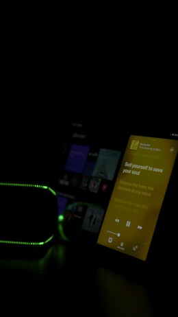 The image features a dimly lit room with two electronic devices visible. One device displays a screen with a music app playing, showing lyrics in yellow against a black backdrop. Another device, possibly a speaker, has a green light outline. The overall lighting is low, emphasizing the glow from the screen and lights.