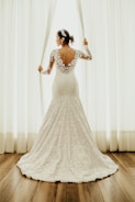 A bride wearing a lace wedding gown stands by a window, holding open white curtains. The dress features a floral lace design with an open back. Her hair is styled with a decorative headpiece, and the setting is enhanced by natural light filtering through the window.