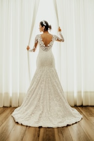 A bride wearing a lace wedding gown stands by a window, holding open white curtains. The dress features a floral lace design with an open back. Her hair is styled with a decorative headpiece, and the setting is enhanced by natural light filtering through the window.