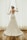 A bride wearing a lace wedding gown stands by a window, holding open white curtains. The dress features a floral lace design with an open back. Her hair is styled with a decorative headpiece, and the setting is enhanced by natural light filtering through the window.