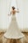 A bride wearing a lace wedding gown stands by a window, holding open white curtains. The dress features a floral lace design with an open back. Her hair is styled with a decorative headpiece, and the setting is enhanced by natural light filtering through the window.