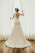 A bride wearing a lace wedding gown stands by a window, holding open white curtains. The dress features a floral lace design with an open back. Her hair is styled with a decorative headpiece, and the setting is enhanced by natural light filtering through the window.