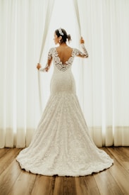 A bride wearing a lace wedding gown stands by a window, holding open white curtains. The dress features a floral lace design with an open back. Her hair is styled with a decorative headpiece, and the setting is enhanced by natural light filtering through the window.